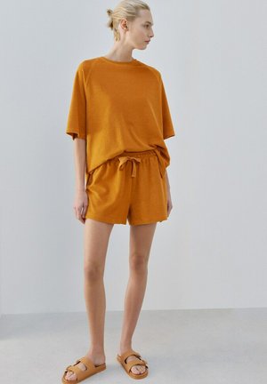 Donna con capelli biondi raccolti in uno chignon, indossa una maglietta arancione a maniche corte, pantaloncini abbinati con coulisse e sandali beige a ciabatta, in piedi all'interno.