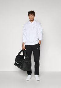 Weißer Baumwoll-Hoodie mit Logo, schwarze Jogginghose mit Logo, hält eine schwarze Sporttasche mit Netzseite und weiße Sneaker. Schlichte, lässige Kleidung.
