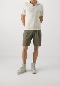 Polo shirt beige a maniche corte con scollo a V, abbinato a pantaloni corti verde oliva. Sneakers alte grigie completano l'outfit.