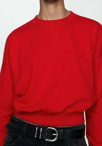 Person trägt ein knallrotes Sweatshirt, das in schwarze Hosen mit einem schwarzen Gürtel und silberner Schnalle gesteckt ist, zu sehen vom Hals bis zu den Hüften.