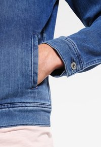 Chaqueta de denim con un tejido azul texturizado. Las características incluyen un puño ajustado con botón a presión y un bolsillo lateral.