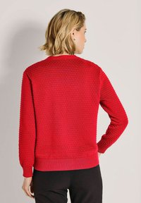 Frau mit kurzem blonden Haar, die einen strukturierten roten Pullover und schwarze Hosen trägt, mit dem Rücken zugewandt und einer Hand auf der Hüfte.