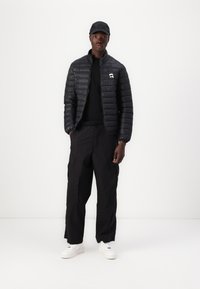 Schwarze Steppjacke mit horizontaler Quiltung, einem Frontreißverschluss und Logodetail. Kombiniert mit schwarzen Cargo-Hosen und weißen Turnschuhen.