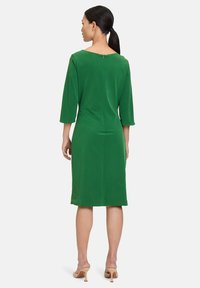 Grünes knielanges Kleid mit ¾ Ärmeln, einem Boot-Ausschnitt und einem Reißverschluss auf der Rückseite. Glatte Textur und minimalistisches Design.
