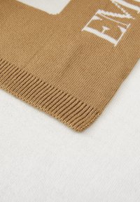 Sciarpa lavorata a maglia color beige con bordi a coste. Presenta scritte bianche e una trama liscia su uno sfondo bianco.