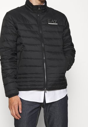 Homme portant une veste matelassée noire EA7 sur une chemise blanche et un jean foncé, les mains détendues — veste zippée avec logo sur la poitrine.