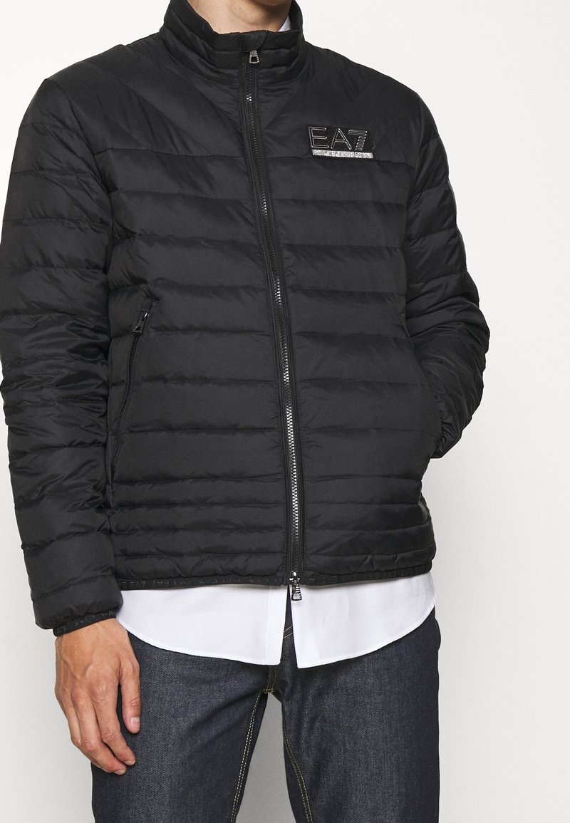 Homme portant une veste matelassée noire EA7 sur une chemise blanche et un jean foncé, les mains détendues — veste zippée avec logo sur la poitrine.