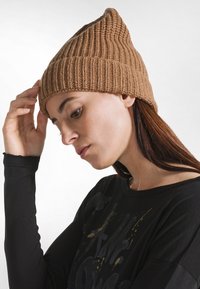 Beanie lavorato a maglia in marrone chiaro con una texture a coste e una corona appuntita. Abbinato a un top a maniche lunghe nero con accenti grafici.