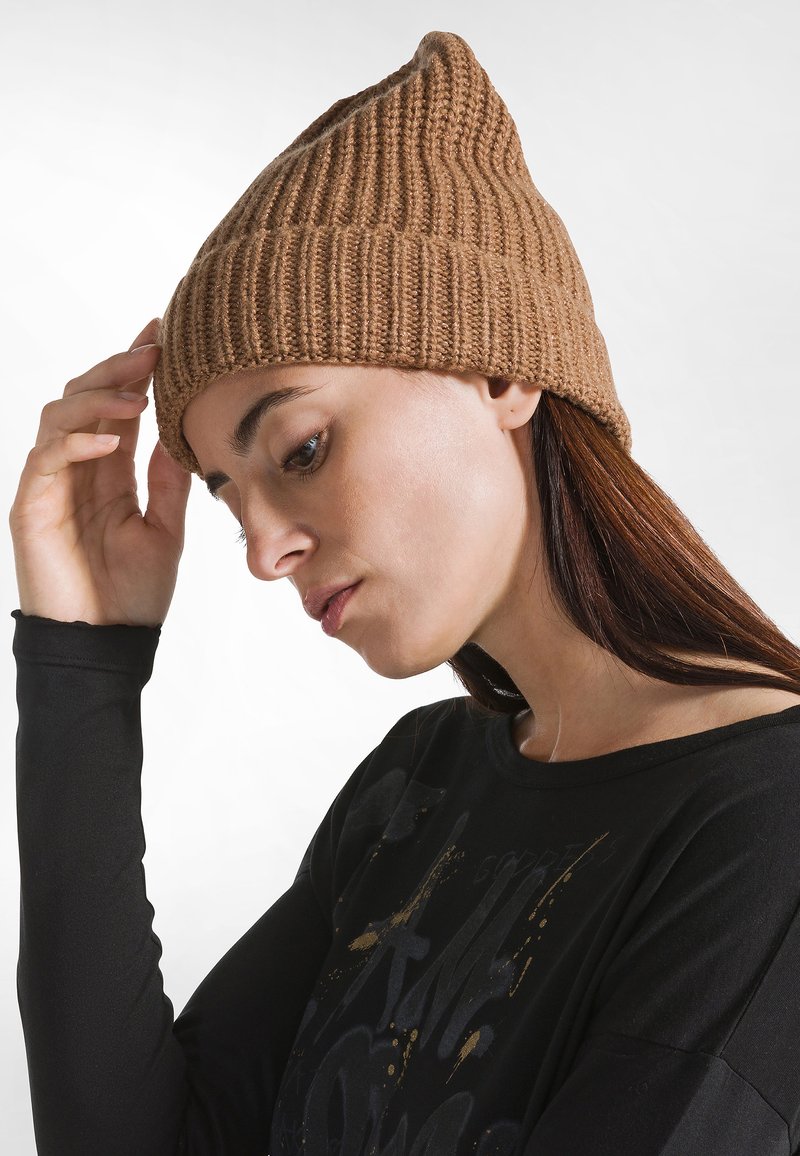 Beanie lavorato a maglia in marrone chiaro con una texture a coste e una corona appuntita. Abbinato a un top a maniche lunghe nero con accenti grafici.