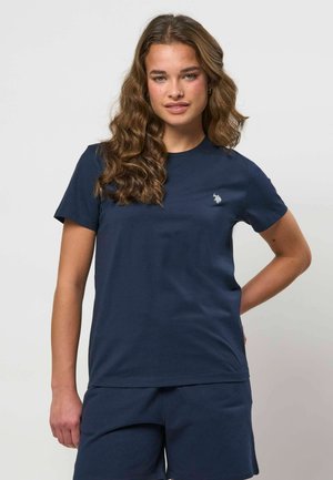 Žena s dlhými vlnitými vlasmi, oblečená v námornícky modrom tričku s malým bielym vyšitým logom a zodpovedajúcich námornícky modrých šortkách.