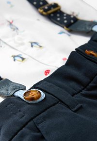 Gros plan sur un pantalon bleu foncé avec un bouton marron et une boucle de bouton tressée bleue, une chemise blanche avec de petits motifs pingouin et champignon rouge, et des bretelles.