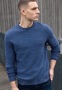 Jersey azul de punto con cuello redondo, tejido texturizado y mangas largas. Combinado con vaqueros oscuros que muestran un diseño ajustado.