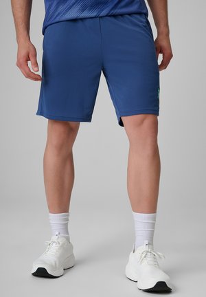 Mężczyzna w niebieskich sportowych szortach, białych skarpetach do kostki i białych butach do biegania stojący na jednolitej szarej powierzchni.
