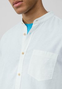 Witte linnen shirt met een bandkraag, twee knopen bij de hals en een borstzak. Beschikt over lichte houten knopen en een zachte textuur.