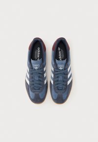 Zapatillas deportivas de ante azul con rayas blancas, acentos en color burdeos y suela de goma. Punta redonda y diseño con cordones. Aspecto limpio y clásico.