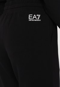 Fekete sportnadrág puha anyagból, elasztikus derékkal. Az oldalon fehér EA7 Emporio Armani logó található.