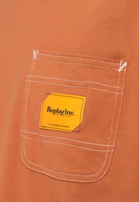 Πορτοκαλί υφασμάτινη τσέπη με λευκή ραφή και μια κίτρινη ετικέτα που γράφει «Replay Inc. Engineered Works» ραμμένη πάνω της.