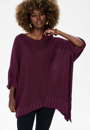 Vrouw met krullend haar, gouden oorringen en armband, gekleed in een losse paarse gebreide trui en zwarte broek, staand tegen een witte achtergrond.