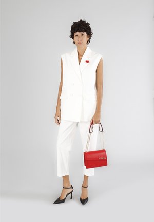 Blazer bianco senza maniche con bottoni doppi, abbinato a pantaloni bianchi su misura. Tacchi a punta neri e una borsa rossa con tracolla a catena.