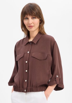 Frau mit braunen Haaren, die eine lockere braune Jacke mit Knöpfen und Klappentaschen sowie weiße Hosen trägt, steht vor einem einfarbigen hellen Hintergrund.