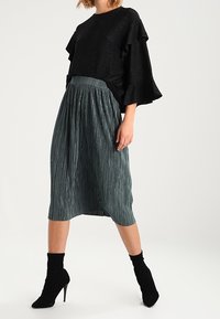 Top negro con mangas acampanadas y textura brillante, combinado con una falda midi de plisados en color teal. Llevado con botas de tacón alto negras hasta el tobillo.