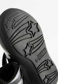 Schwarze Schuhsohle mit einem sternförmigen Profil, das Rillen und Linien für Grip aufweist. Der Zehenbereich ist nach oben gebogen. Das Etikett sagt "NEXT."