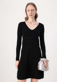 Black knit dress with long sleeves, V-neck, and ruched waist; features horizontal lines and a flared skirt. Holding a silver satin pouch.