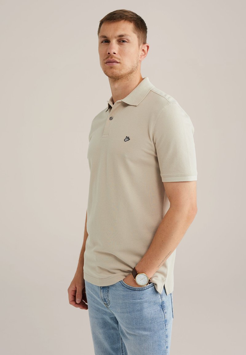 Tricou polo de culoare bej deschis, confectionat dintr-un material texturat, având un guler cu trei nasturi și un mic logo negru pe piept. Purtați-l cu blugi de culoare albastră deschis.