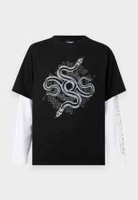 Μαύρο πουκάμισο με κοντά μανίκια φορεμένο πάνω από λευκά μακριά μανίκια, με μεγάλο λευκό γραφικό παράσταση ενός πλεγμένου φιδιού και στυλιζαρισμένο κείμενο στο μπροστινό μέρος.