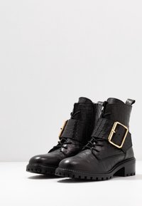 Botas de tobillo de cuero texturizado negro con suelas gruesas, cremalleras laterales, cordones y grandes hebillas doradas sobre una superficie blanca.