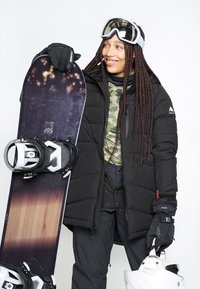 Zwarte pufferjack, camouflage top en donkere sneeuwbroek. Houdt een donkere snowboard met witte bindingen vast en heeft een helm in de andere hand.