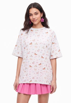 Ung kvinde iført hvid oversized T-shirt med små farverige gyngehest prints og en lyserød plisseret mininederdel, smilende til kameraet.