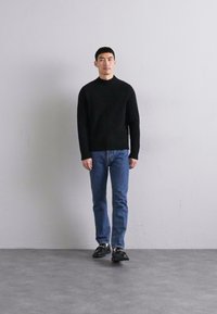 Μαύρο πλεκτό πουλόβερ με ψηλό γιακά, μπλε τζιν και μαύρα παπούτσια, σκύβοντας μπροστά σε έναν ανοιχτό γκρίζο τοίχο με απλό φόντο και ουδέτερο δάπεδο.