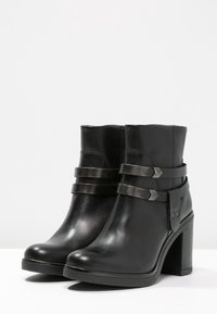 Botas de cuero negras hasta el tobillo con un tacón grueso, que cuentan con dos correas horizontales con acentos metálicos y una textura suave y brillante.