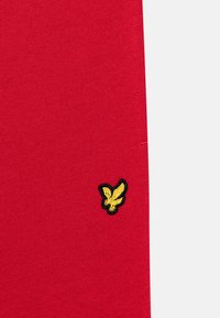 Tissu tricoté rouge avec un petit logo d'oiseau brodé en noir et jaune en bas à gauche. Texture lisse avec une couleur unie.