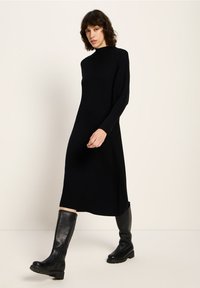 Schwarzes geripptes Strickkleid mit hohem Ausschnitt und langen Ärmeln, das bis zur Mitte des Wades reicht. Kombiniert mit kniehohen schwarzen Lederstiefeln.