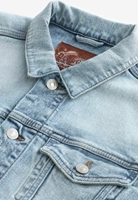 Lichtblauwe spijkerjack met een klassieke kraag, zilveren drukknopen en een leren label. Voorzien van dubbele borstzakken en beige stiksels.