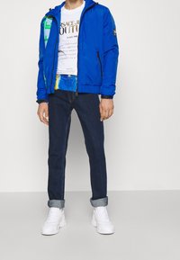 Blå nylon bomberjacka med huva, åtsittande ärmar och dragkedjefickor. Vit grafisk t-shirt och mörk denimjeans med uppvikta ben. Vita sneakers.