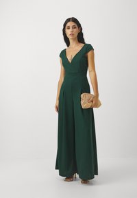 Vihreä haalari, jossa on syvä V-aukko ja hihattomat hihat, minkä lisäksi se on vyötäröltä istuva ja leveälahkeinen. Pitää pientä kultaisesta verkkokangasta valmistettua clutch-laukku.