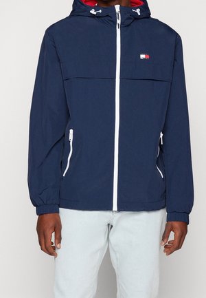 Mężczyzna ubrany w granatową kurtkę z kapturem, białym zamkiem błyskawicznym i kieszeniami, z małym logo Tommy Hilfiger na piersi, w połączeniu z jasnymi jeansami.