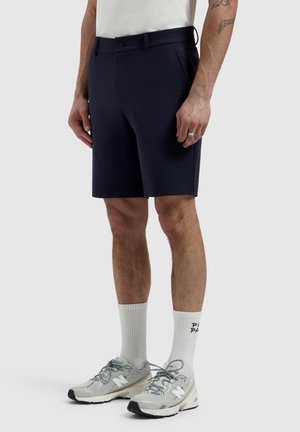 Man met donkerblauwe korte broek, witte sokken met tekst, grijze New Balance sneakers en een wit shirt, staand met ontspannen handen langs het lichaam.