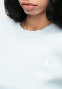 Κοντινότερη λήψη ενός ατόμου που φοράει ένα ανοιχτό μπλε φούτερ με ένα μικρό κεντημένο λευκό λογότυπο Jumpman στο στήθος.