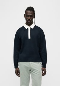 Μπλε μπλουζάκι με λευκό γιακά πόλο, μακριά μανίκια και άνετη εφαρμογή· συνδυάζεται με ανοιχτοπράσινο παντελόνι με υφή.