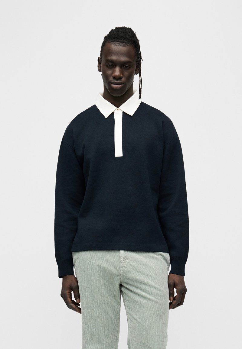 Μπλε μπλουζάκι με λευκό γιακά πόλο, μακριά μανίκια και άνετη εφαρμογή· συνδυάζεται με ανοιχτοπράσινο παντελόνι με υφή.