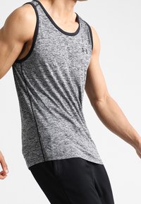 Hombre vistiendo una camiseta atlética sin mangas de color gris y pantalones negros, posando con los brazos ligeramente doblados contra un fondo claro y simple.