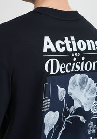 Zwarte sweatshirt met witte gedrukte tekst "Acties en Beslissingen" en bloemgrafieken, met een ronde halslijn en gestructureerd materiaal.
