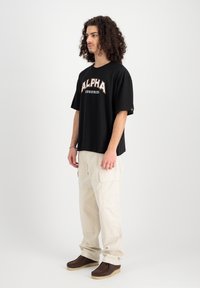 Fekete, oversized póló "ALPHA INDUSTRIES" narancssárga és fehér felirattal, krémszínű cargo nadrággal és barna slip-on cipőkkel párosítva.