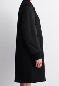 Cappotto nero con maniche lunghe; corpo in lana e maniche imbottite in nylon. Presenta polsini a costine, tasche laterali e una silhouette dritta.
