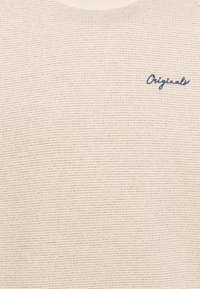 Suéter color beige claro con un patrón de canalé texturizado. Presenta un logo bordado en azul marino que dice "Originals" en el pecho.