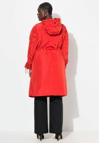 Veste imperméable rouge avec taille à cordon, capuche et poignets élastiques, associée à un pantalon large noir et des talons. Texture lisse, tissu léger.